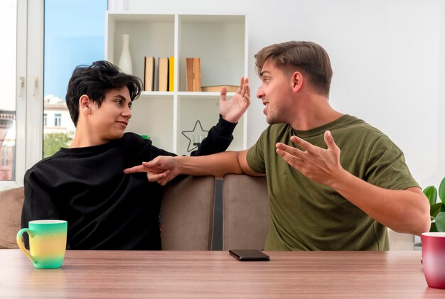 Joyful young blonde handsome man sitting at table looking and pointing at pleased young brunette handsome boy raising hand