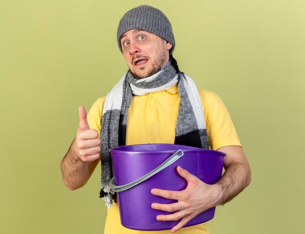 Радостный молодой блондин больной славянский мужчина в зимней шапке и шарфе показывает палец вверх и держит пластиковое ведро, изолированное на оливково-зеленой стене с копией пространства