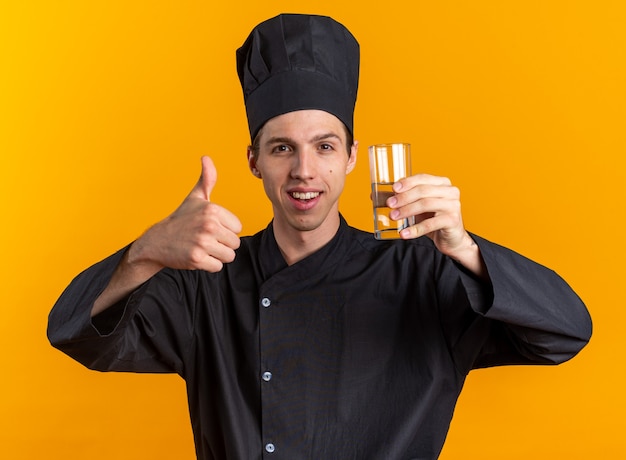 Joyful young blonde male cook in chef uniform and cap holding glass of water showing thumb up