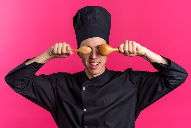 Foto gratuita gioioso giovane maschio biondo cuoco in uniforme da chef e cappello che tiene i cucchiai davanti agli occhi