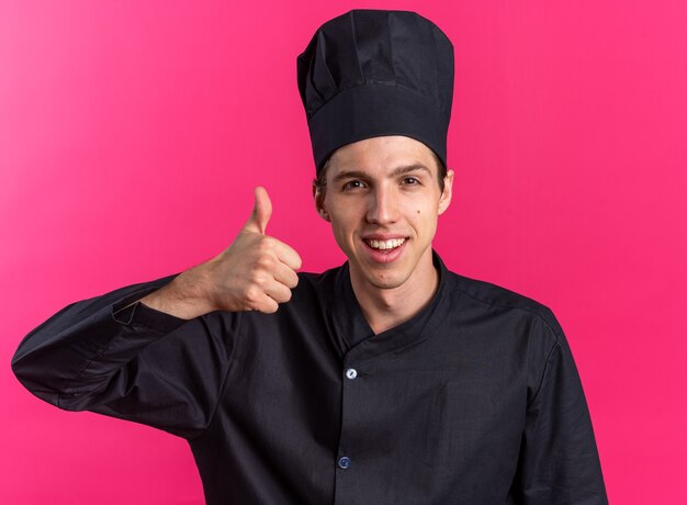 Joyful young blonde male cook in chef uniform and cap showing thumb up 
