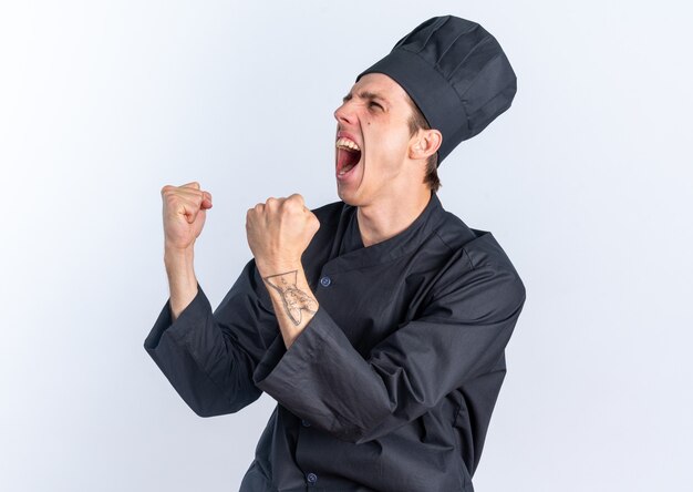 Joyful young blonde male cook in chef uniform and cap standing in profile view looking at side doing yes gesture 