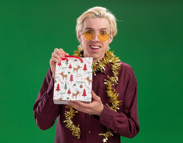 Joyful young blonde man wearing glasses with tinsel garland around neck holding christmas gift bag  isolated on green wall
