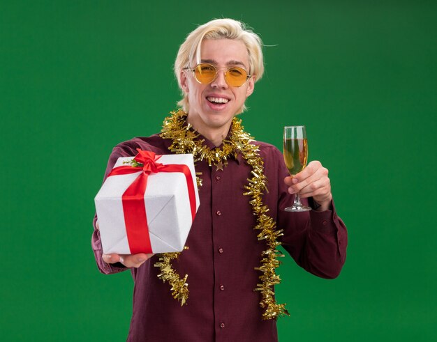 Joyful young blonde man wearing glasses with tinsel garland around neck holding glass of champagne and stretching out gift package   isolated on green wall