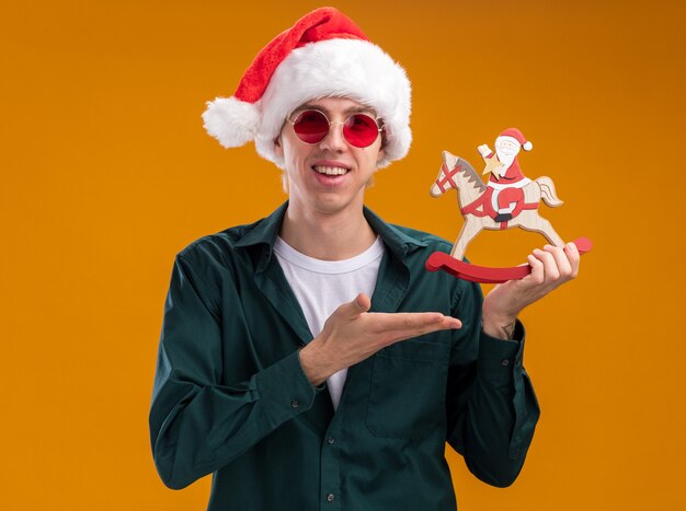 Gioioso giovane biondo che indossa cappello da babbo natale e occhiali che tiene e indica con la mano a santa sulla figurina di cavallo a dondolo guardando la telecamera isolata su sfondo arancione