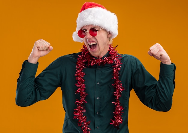 Joyful young blonde man wearing santa hat and glasses with tinsel garland around neck doing yes gesture with closed eyes isolated on orange background