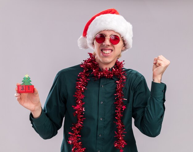 Joyful young blonde man wearing santa hat and glasses with tinsel garland around neck holding christmas tree toy with date looking at camera doing yes gesture winking isolated on white background