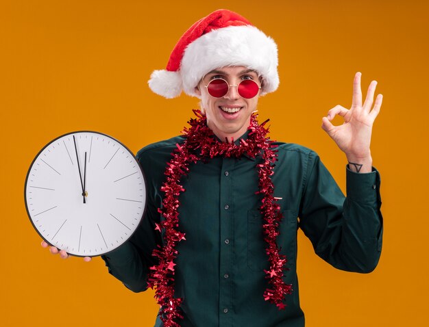 오렌지 배경에 고립 된 확인 서명을하고 카메라를보고 시계를 들고 목 주위에 반짝이 갈 랜드와 산타 모자와 안경을 쓰고 즐거운 젊은 금발의 남자