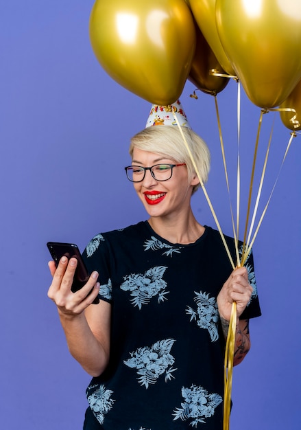 Радостная молодая блондинка тусовщица в очках и кепке дня рождения держит воздушные шары и мобильный телефон, глядя на телефон, изолированные на фиолетовом фоне