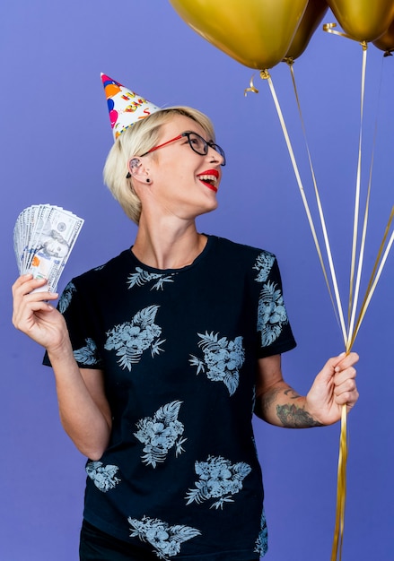 Joyful young blonde party girl wearing glasses and birthday cap holding balloons and money looking at side isolated on purple background