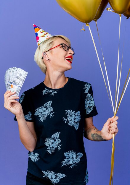 紫色の背景で隔離の側を見て風船とお金を保持している眼鏡と誕生日の帽子を身に着けているうれしそうな若いブロンドのパーティーの女の子