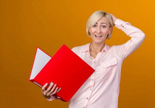 Joyful young blonde slavic woman holding folder and putting hand on behind head isolated on orange wall