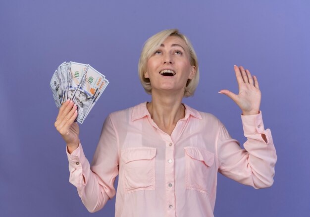 Радостная молодая белокурая славянская женщина смотрит вверх, держа деньги и показывая пустую руку, изолированную на фиолетовом фоне