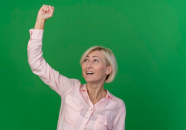 Радостная молодая блондинка славянская женщина поднимает кулак и смотрит на него изолированно на зеленом фоне с копией пространства