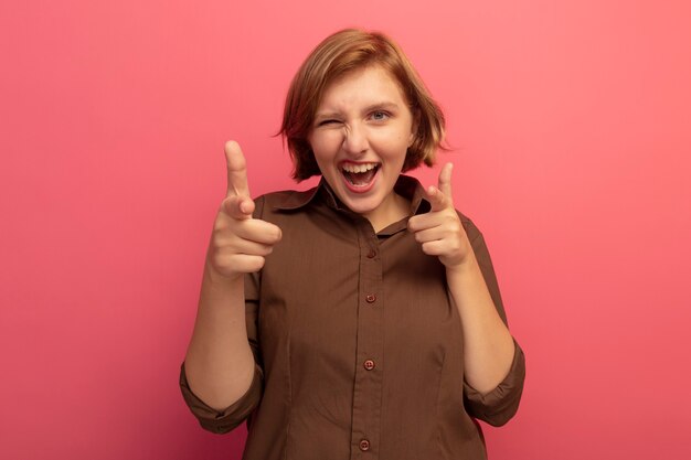 Foto gratuita gioiosa giovane donna bionda che guarda l'occhiolino anteriore facendo un gesto isolato sulla parete rosa con spazio per le copie