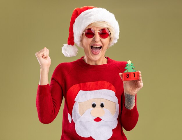 joyful young blonde woman wearing christmas hat and santa claus christmas sweater with glasses holding christmas tree toy with date doing yes gesture with closed eyes isolated on olive green wall