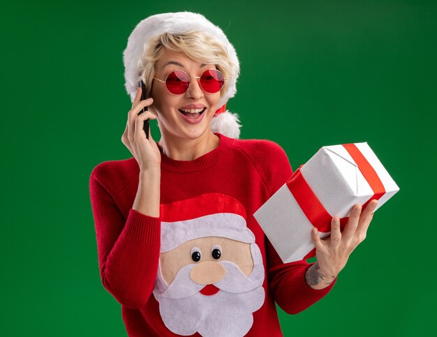 Joyful young blonde woman wearing christmas hat and santa claus christmas sweater with glasses holding and looking at gift package talking on phone isolated on green wall