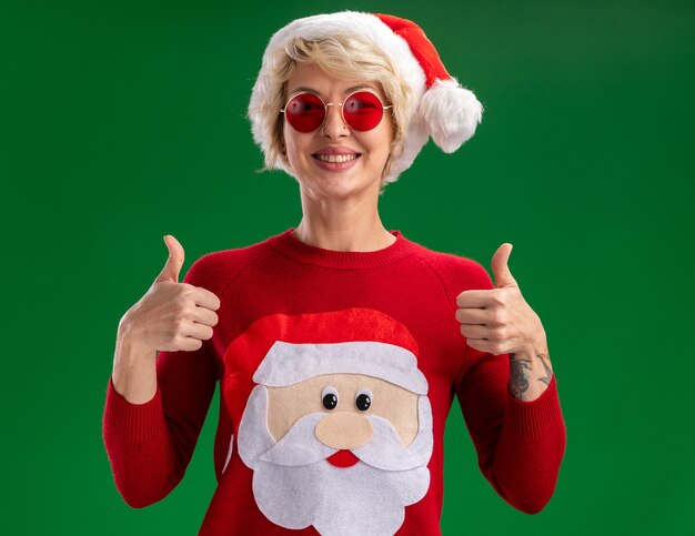 クリスマスの帽子とサンタクロースのクリスマスセーターを身に着けているうれしそうな若いブロンドの女性は、緑の背景に分離された親指を示すカメラを見てメガネ