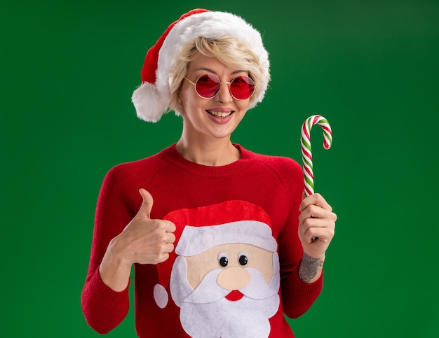 크리스마스 모자와 산타 클로스 크리스마스 스웨터를 입고 즐거운 젊은 금발의 여자는 녹색 벽에 고립 된 엄지 손가락을 보여주는 크리스마스 사탕 지팡이를 들고 찾고 안경