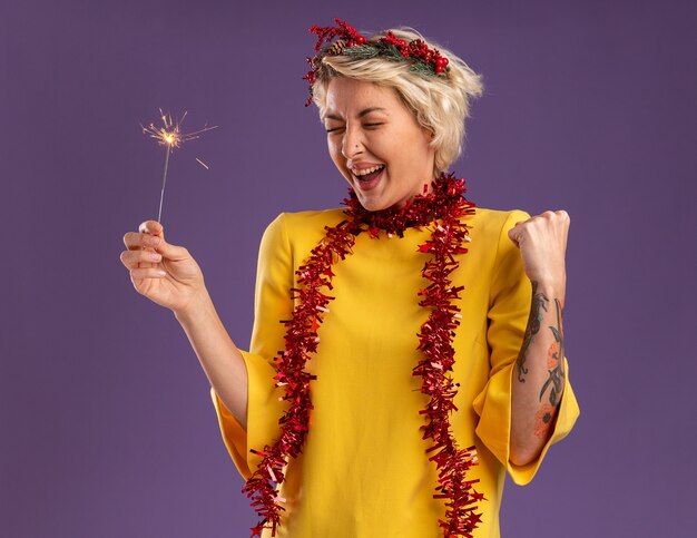 Радостная молодая блондинка в рождественском венке и гирлянде из мишуры на шее держит праздничный бенгальский огонь, делая жест "да" с закрытыми глазами, изолированными на фиолетовой стене