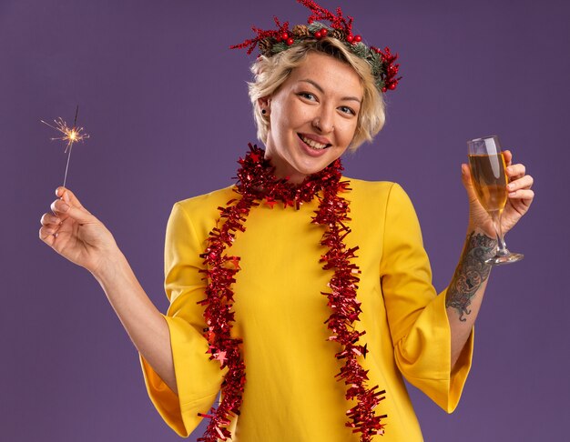 Радостная молодая блондинка в рождественском венке и гирлянде из мишуры на шее держит праздничный бенгальский огонь и бокал шампанского, изолированные на фиолетовой стене
