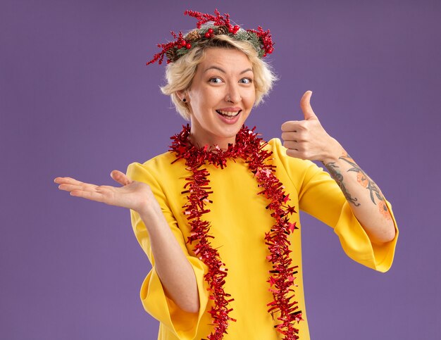 Joyful young blonde woman wearing christmas head wreath and tinsel garland around neck looking  showing empty hand and thumb up isolated on purple wall