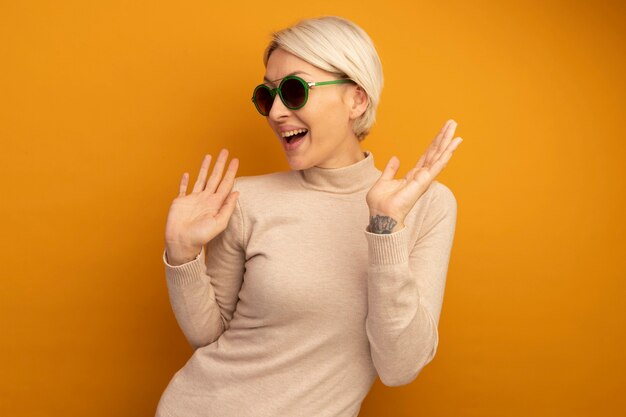 Joyful young blonde woman wearing sunglasses looking at side showing empty hands isolated on orange wall