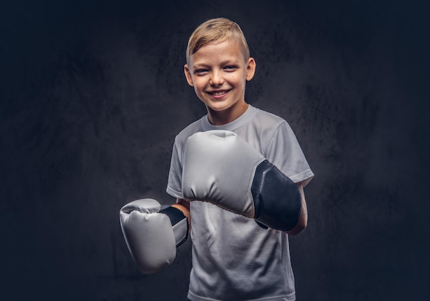 Joyful young boy boxer with blonde hair dressed in a white t-shirt with boxing gloves posing in a studio. Isolated on the dark textured background.