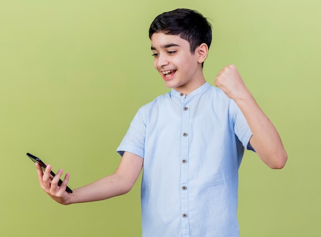 Joyful young boy holding and looking at mobile phone doing yes gesture isolated on olive green wall