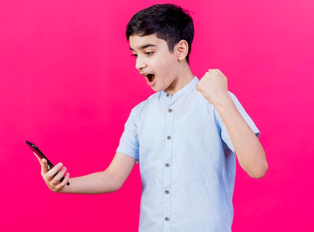 Радостный мальчик, держащий и смотрящий на мобильный телефон, делает жест да, изолированный на розовой стене