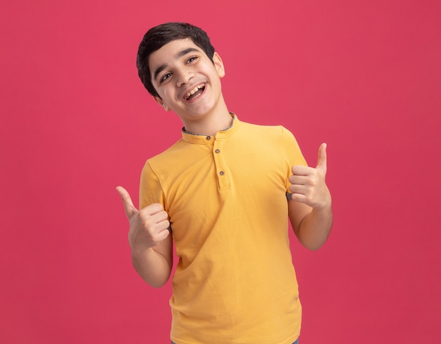 Joyful young boy looking at side showing thumbs up isolated on pink wall