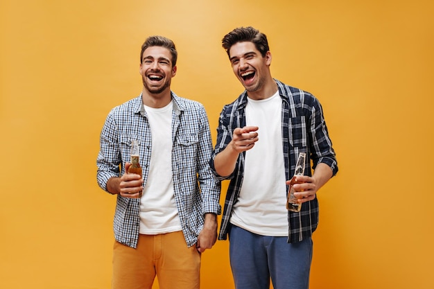화려한 반바지와 체크무늬 셔츠를 입은 유쾌한 젊은 브루넷 남성들은 카메라를 가리키며 오렌지색 배경에 맥주병을 들고 있다