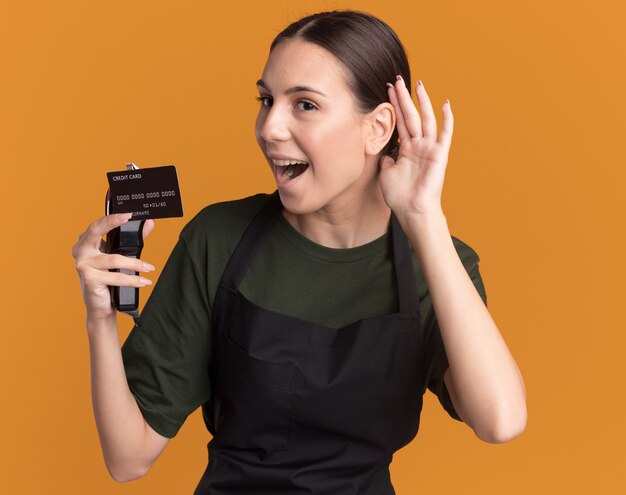 Joyful young brunette barber girl in uniform holds hair clippers and credit card keeping hand behind ear