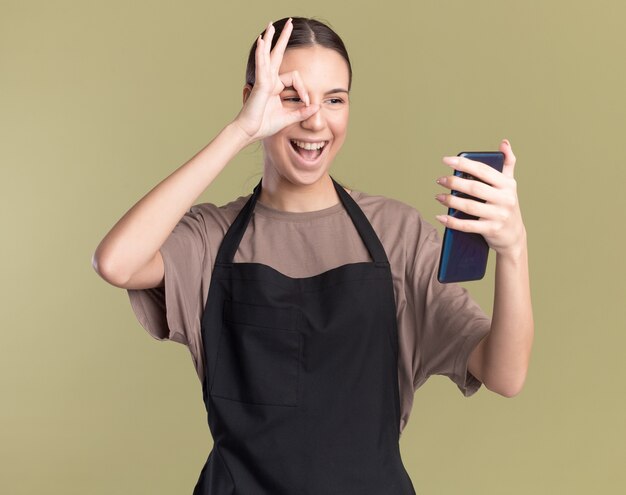 Joyful young brunette barber girl in uniform holds and looks at phone through fingers on olive green