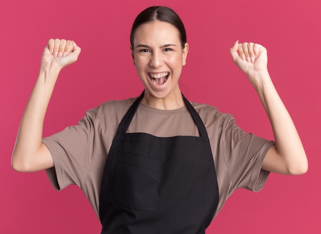 Foto gratuita gioiosa giovane ragazza bruna barbiere in uniforme si leva in piedi con i pugni alzati isolati sulla parete rosa con lo spazio della copia