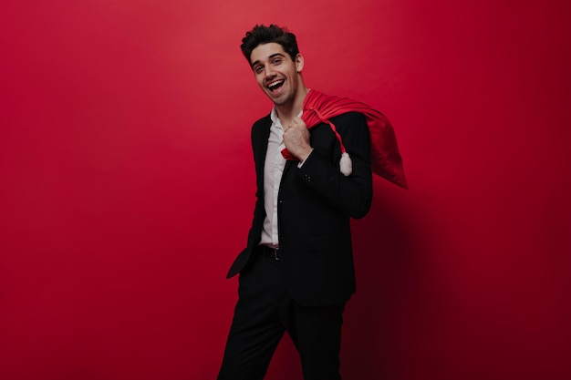 Joyful young brunette in black suit white shirt posing with bright Santa Claus bag looking into camera isolated on red background