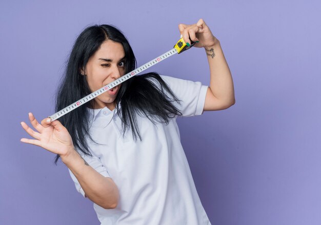 Joyful young brunette caucasian girl blinks eye and holds tape measure isolated on purple wall