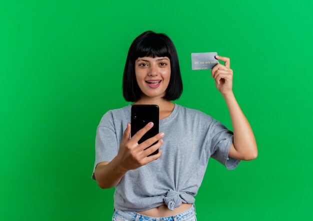 うれしそうな若いブルネット白人の女の子はクレジットカードと電話を保持します