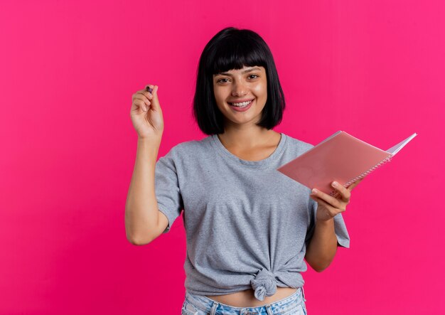 Joyful young brunette caucasian girl holds pen and notebook looking  