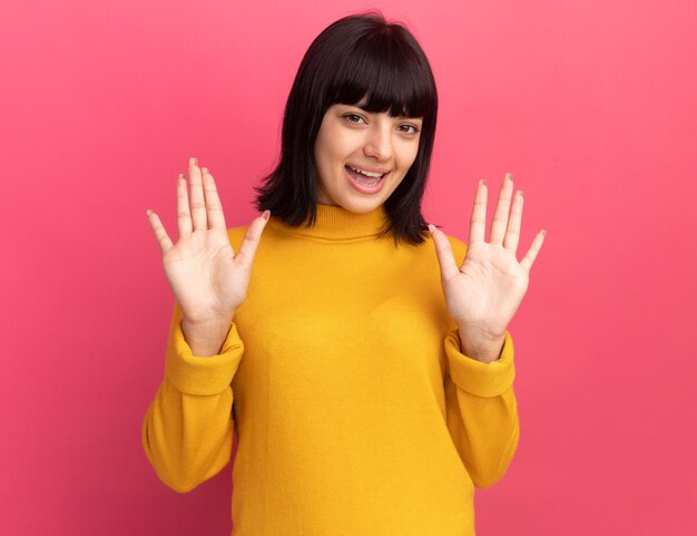 Joyful young brunette caucasian girl stands with raised hands on pink