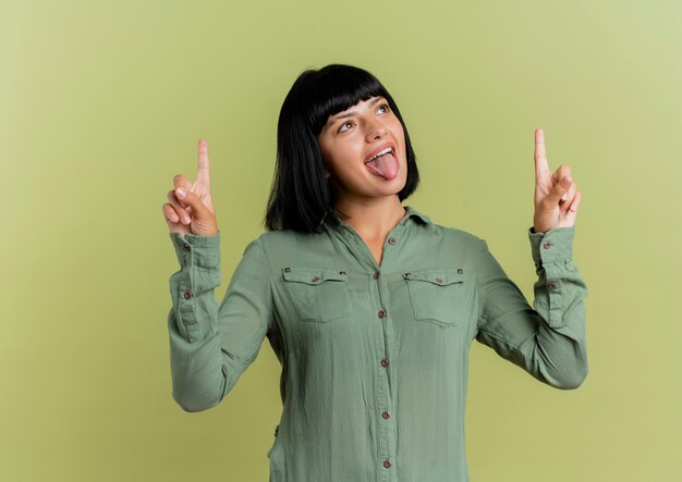 Joyful young brunette caucasian girl stucks out tongue and points up with two hands isolated on olive green background with copy space