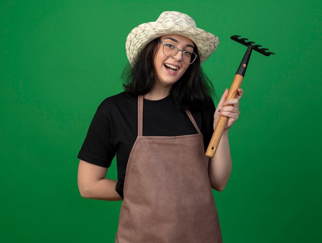 Joyful young brunette female gardener in optical glasses and uniform wearing gardening hat holds rake and keeps hand behind back isolated on green wall