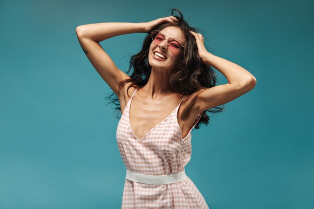 Joyful young brunette with beautiful wavy hairstyle in pink glasses and summer light outfit playing her hair on blue wall