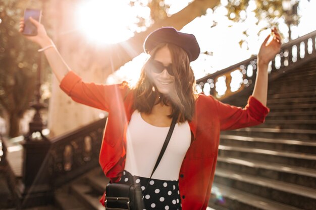 Joyful young brunette with beret stylish white top red shirt and black sunglasses walking in sunny city smiling and looking away against old stairs background