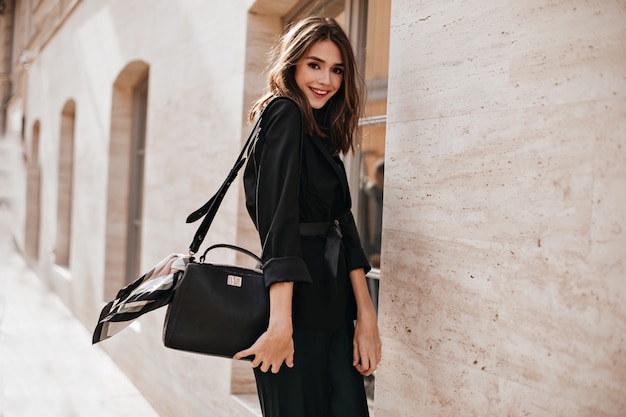 Joyful young brunette with fluffy hair, red lips, trendy dress and black jacket, belt on waist standing in profile at sunny street and smiling against light building wall