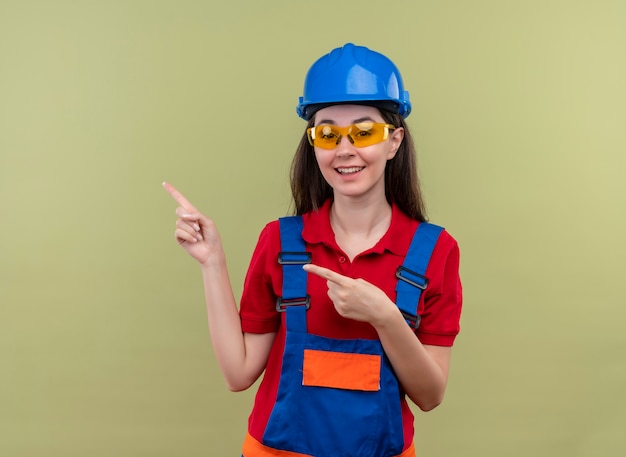 Free photo joyful young builder girl with blue safety helmet and with safety glasses points to the side with both hands on isolated green background with copy space