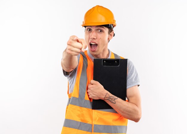 Joyful young builder man in uniform holding clipboard showing you gesture 