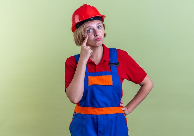 オリーブグリーンの壁に隔離された指でまぶたを引き下げる制服を着たうれしそうな若いビルダーの女性