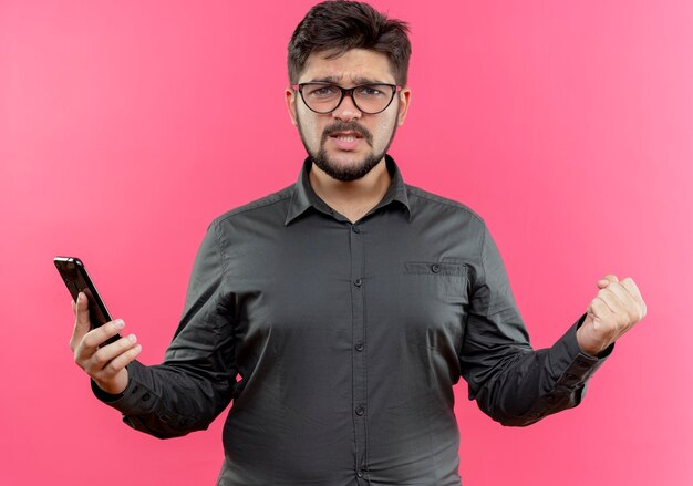 Joyful young businessman wearing glasses holding phone showing yes gesture isolated on pink wall