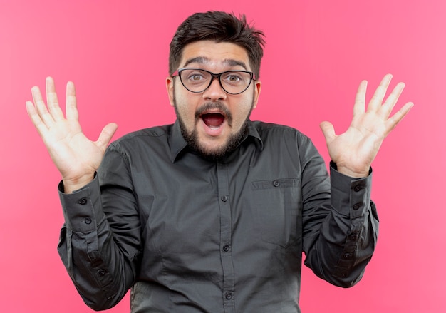 Joyful young businessman wearing glasses spreads hands isolated on pink wall